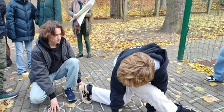 Powiększ grafikę: Uczniowie na placu przy szkole grają w grę ruchową. Na pierwszym planie dwóch chłopców: jeden kuca, a drugi stoi w bardzo dużym wykroku i podpiera się rekami.