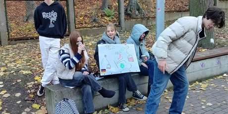 Powiększ grafikę: Uczniowie siedzą na murku przy szkole. W rękach trzymają plakat z napisem TEE i rysunkami akcesoriów niezbędnych do zaparzenia herbaty.