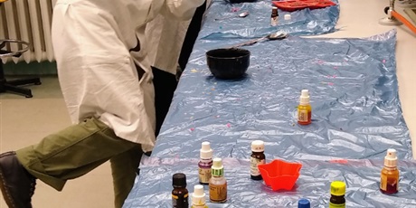 Powiększ grafikę: Zdjęcie stołu laboratoryjnego zabezpieczonego folią. Na stole buteleczki z barwnikami. Obok stoły stoją dwie uśmiechnięte uczennice, które pokazują kciuki w górę.