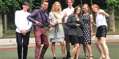 Powiększ grafikę: Zdjęcie grupy uczniów na boisku przed szkołą. Wszyscy są elegancko ubrani. Jedna uczennica trzyma w ręku kwiaty.