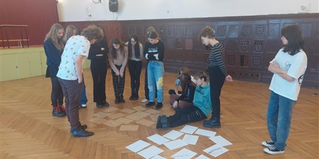 Powiększ grafikę: Grupa uczniów i prowadzący pochylają się nad rozłożonymi na podłodze szarymi i białymi kartkami.