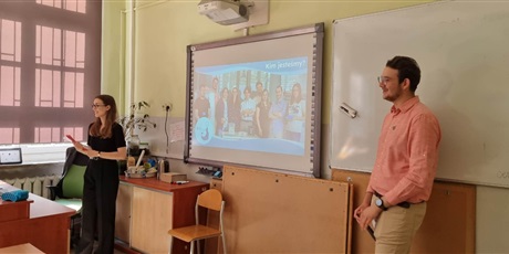Powiększ grafikę: W sali lekcyjnej dwoje prowadzących stoi przy tablicy, na której wyświetlona jest prezentacja multimedialna.
