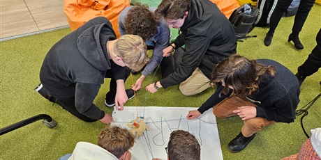 Powiększ grafikę: Grupa uczniów pisze na dużej kartce imię kolegi. Uczniowie piszą grupowo trzymając za sznurki przyczepione do mazaka.