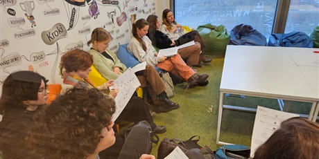 Powiększ grafikę: Grupa uczennic siedzi na pufach i czyta materiały otrzymane na kartkach.