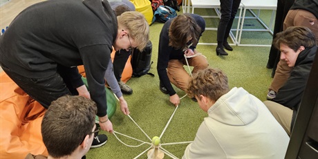 Powiększ grafikę: Uczniowie pracują w grupie próbując przenieść piłeczkę z jednego kołka na drugi trzymając za sznurki przywiązane do podstawki.