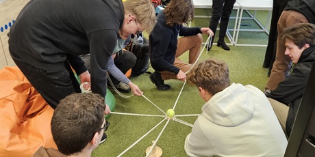 Powiększ grafikę: Uczniowie pracują w grupie próbując przenieść piłeczkę z jednego kołka na drugi trzymając za sznurki przywiązane do podstawki.