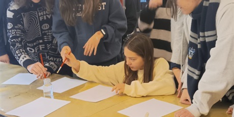 Powiększ grafikę: Grupa uczniów stoi przy stole, na którym leżą białe kartki. Jedna uczennica kuca i zaczyna malować coś pędzlem.