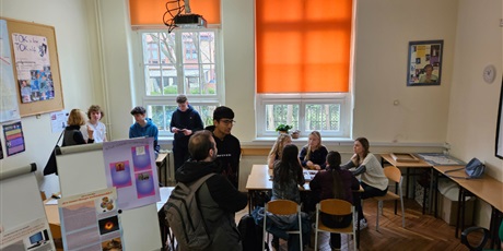 Powiększ grafikę: W sali lekcyjnej stoją flipcharty z plakatami promującymi TOK. Grupa uczennic przy stoliku wyjaśnia coś kandydatom.