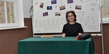 Powiększ grafikę: Uczennica siedzi przy stoliku. Za nią tablica promująca Topomusic