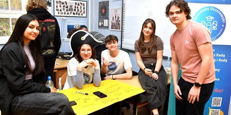 Powiększ grafikę: Grupa uczniów i uczennic przy stoliku, na którym leży duży, żółty arkusz papieru. Obok plakat promujący Parlament Uczniowski.