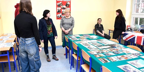 Powiększ grafikę: Grupa uczniów i nauczycielki w sali. Na dużym stole przykrytym zieloną tkaniną wyeksponowane są zdjęcia.