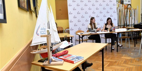 Powiększ grafikę: Stoli, na którym stoi model żaglówki. W tle dwie uczennice siedzące przy stoliku.