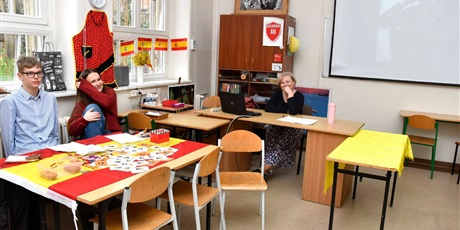 Powiększ grafikę: W sali uczennica i uczeń przy stoliku, na którym leży flaga hiszpańska. Na niej gadżety promujące naukę języka hiszpańskiego.