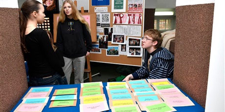 Powiększ grafikę: Dwie uczennice i uczeń za stołem przykrytym niebieską tkaniną, na której leżą kolorowe kartki promujące koła zainteresowań.