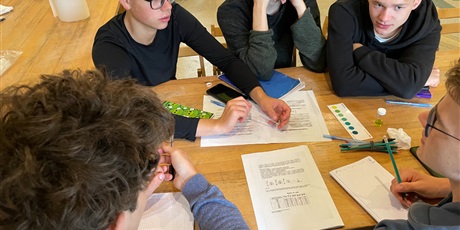 Powiększ grafikę: Zdjęcie grupy uczniów przy stoliku rozwiązujących zadania z chemii.