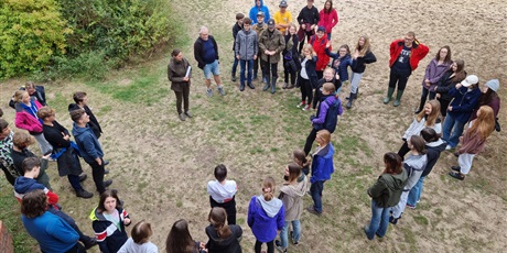 Powiększ grafikę: Zdjęcie dużej grupy uczniów stojących w kręgu i słuchających pani leśnik. Zdjęcie zrobione z góry.