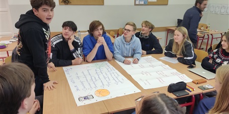 Powiększ grafikę: Grupa uczniów siedząca wokół stołu. Na stale duże arkusze papieru. Jeden uczeń stoi.
