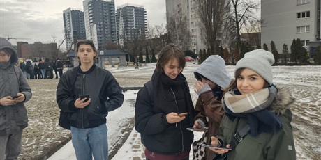 Powiększ grafikę: Grupa uczniów instalujących na telefonach aplikację przy Pomniku Poległych Stoczniowców