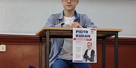 Powiększ grafikę: Uczeń siedzi przy stoliku. Przed nim plakat wyborczy z napisem "Piotr Kudan".