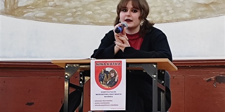 Powiększ grafikę: Uczennica przemawia do mikrofonu siedząc przy stoliku. Przed nią plakat wyborczy z napisem "Nina Katka".