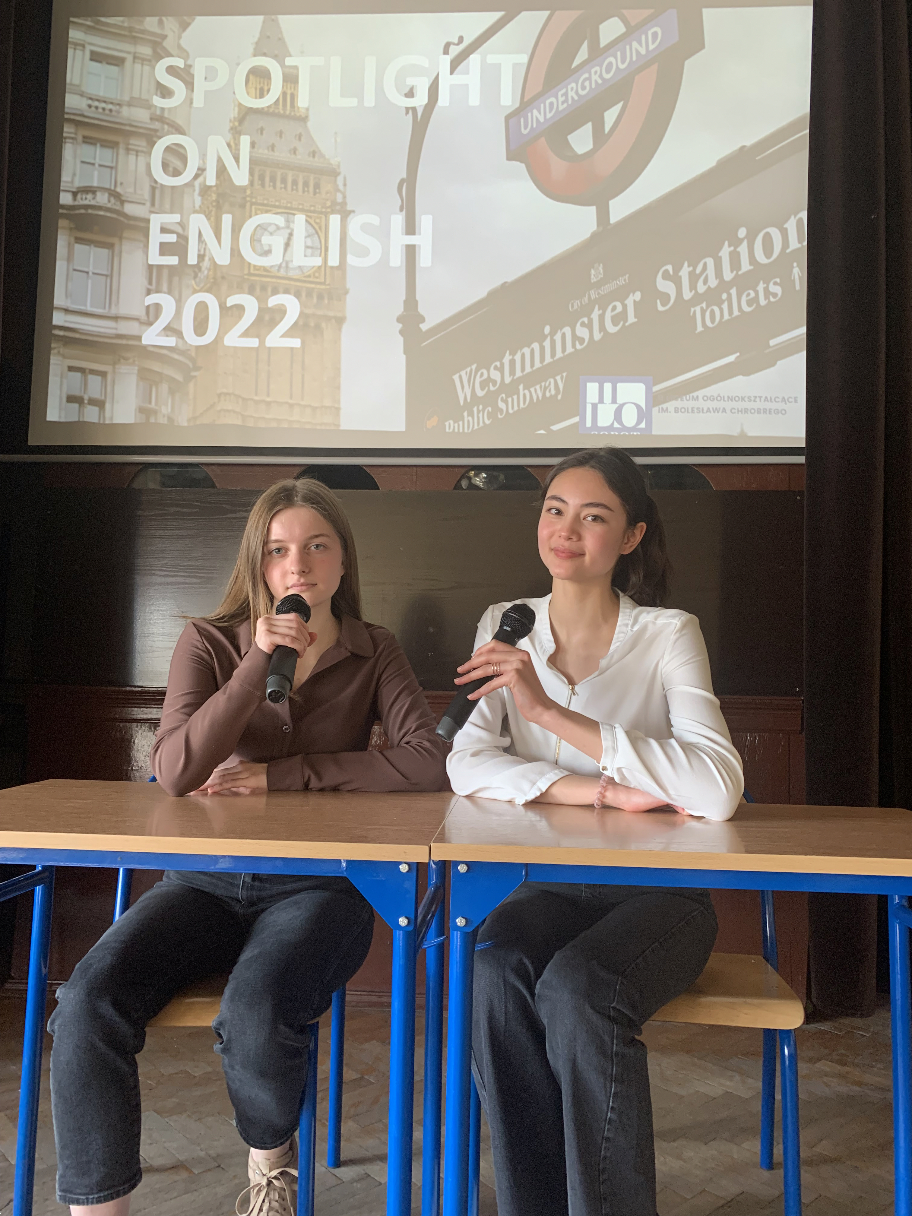 Dwie uczennice w trakcie prezentacji siedzące przy ławce szkolnej, trzymające mikrofony.