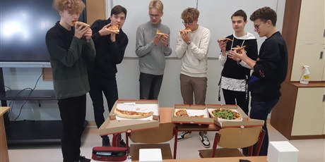 Powiększ grafikę: Zdjęcie 6 uczniów stojących w sali lekcyjnej i jedzących pizzę. Na pierwszym planie leżące na ławce szkolnej kartony z pizzą.